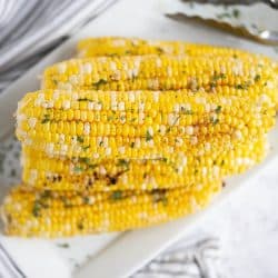 6 ears of grilled corn on the cob stacked on a white rectangle platter. A pair of tongs is in the background.