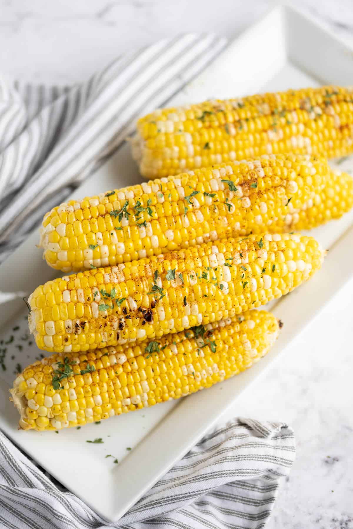 Grilled corn on the cob stacked on a rectangle white platter. Garnished with parsley.