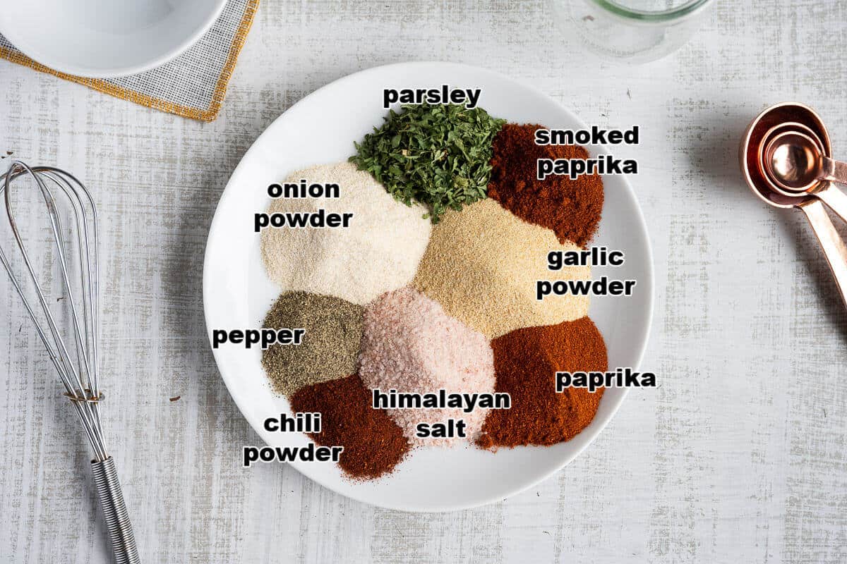 Small white plate with dried spices on it. Spices: parsley, smoked paprika, garlic powder, paprika, salt, chili powder, pepper, and onion powder.