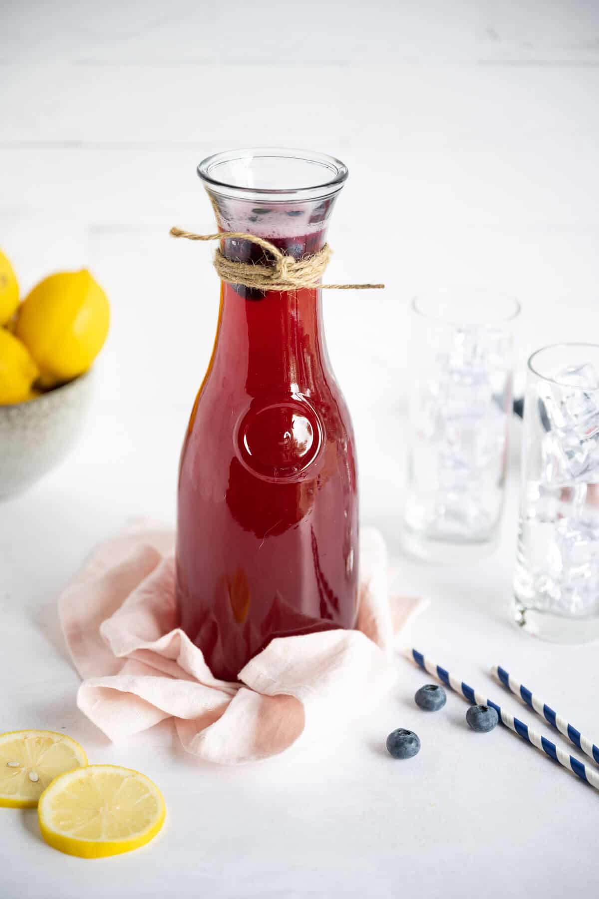 A carafe of homemade blueberry lemonade. Glasses of ice and fresh sliced lemons are nearby.