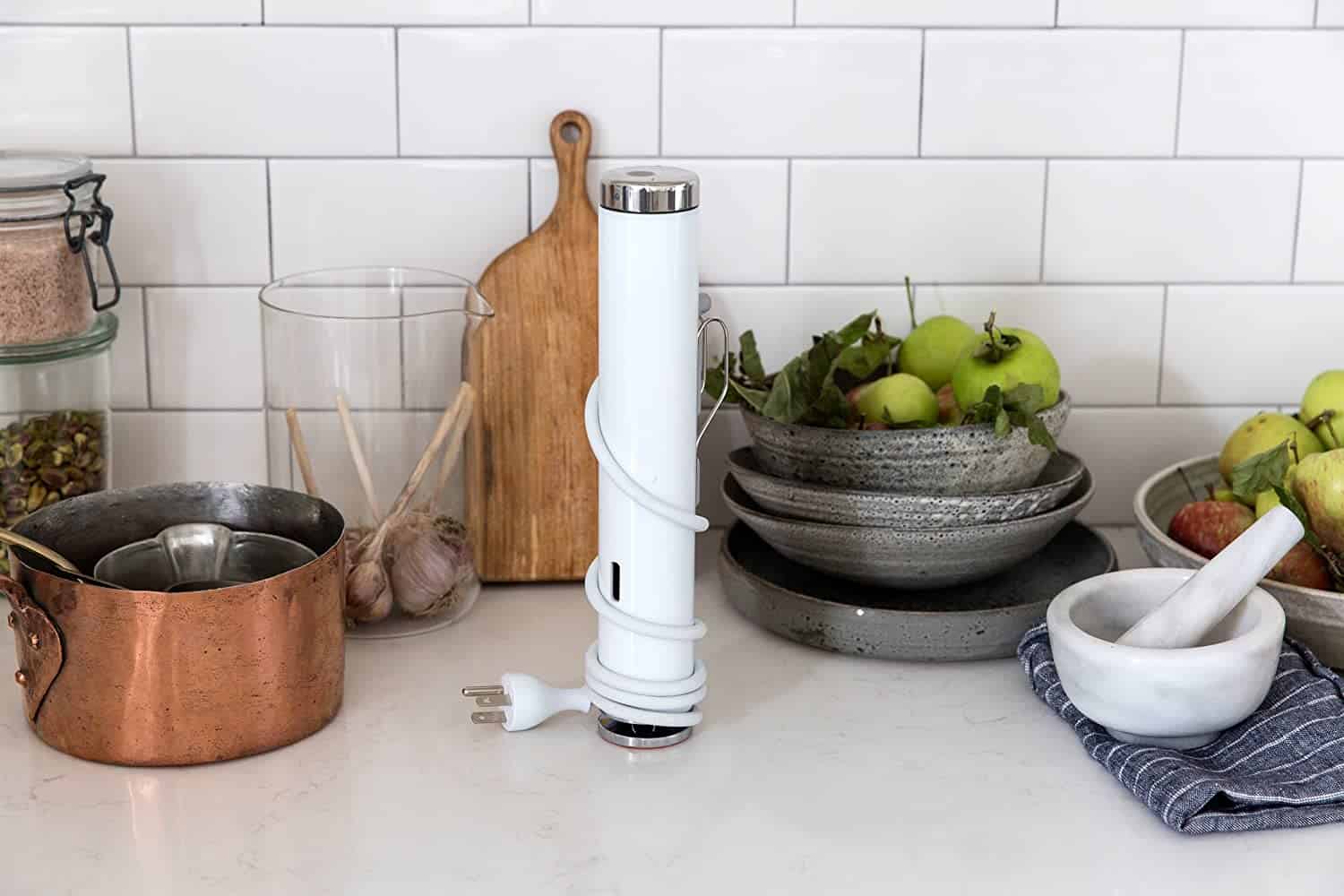 white sous vide wand on the countertop next to some copper pans and metal bowls and a cutting board standing upright against the backsplash