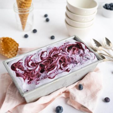 Homemade blueberry ice cream in a loaf pan. Bowls, spoons and Cones are surrounding the ice cream.