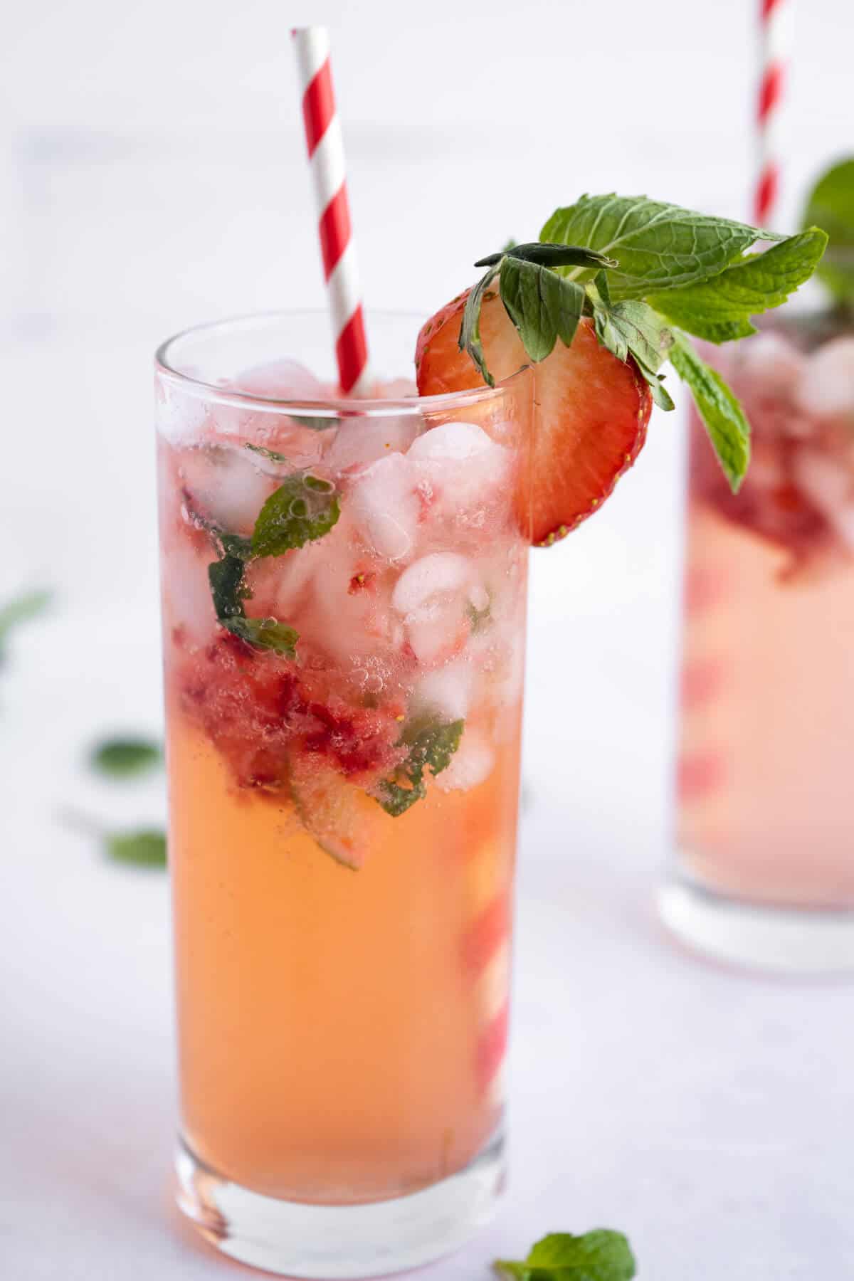 Strawberry Mojito Mocktail in a tall clear glass. Muddled strawberries and mint are visible, drink is finished with a sliced strawberry, fresh mint, and a red and white striped straw.