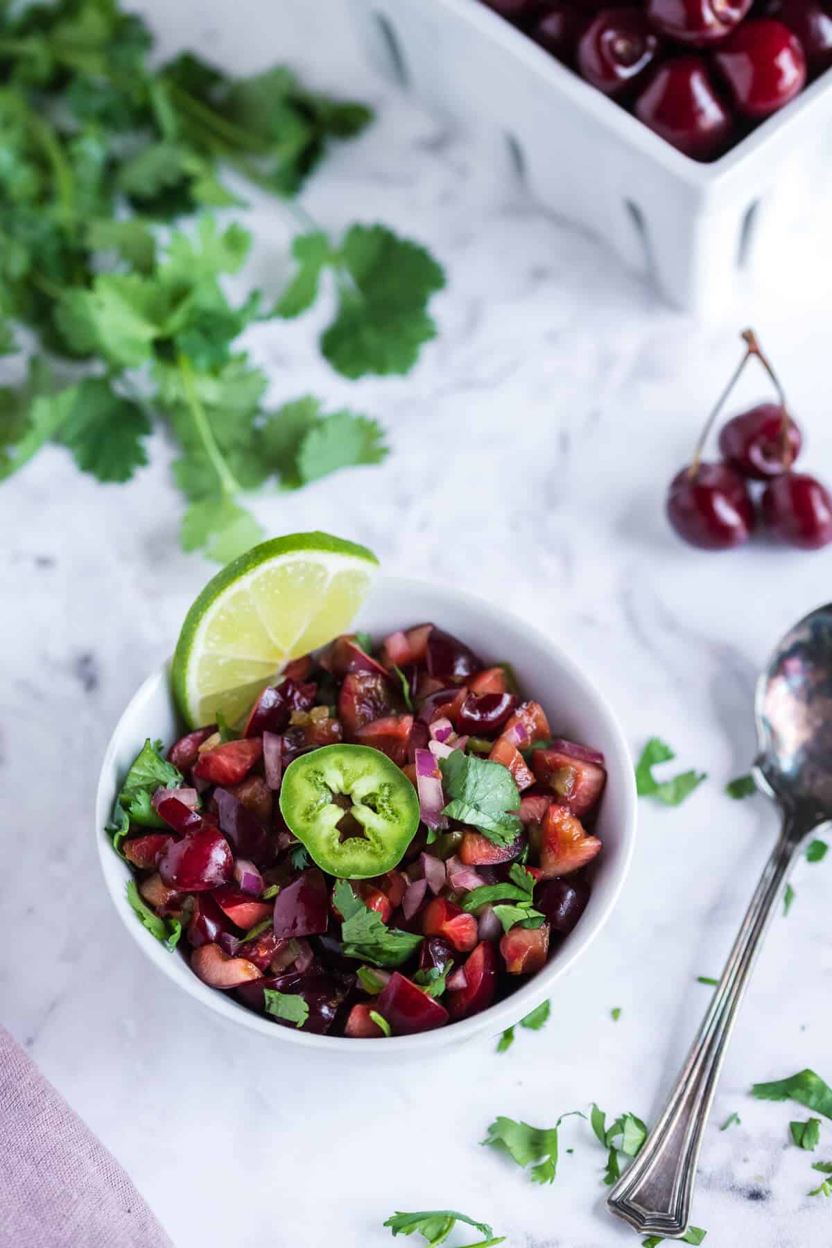 White bowl of fresh cherry salsa garnished with cilantro and a slice of jalapeno. Spoon alongside the bowl.