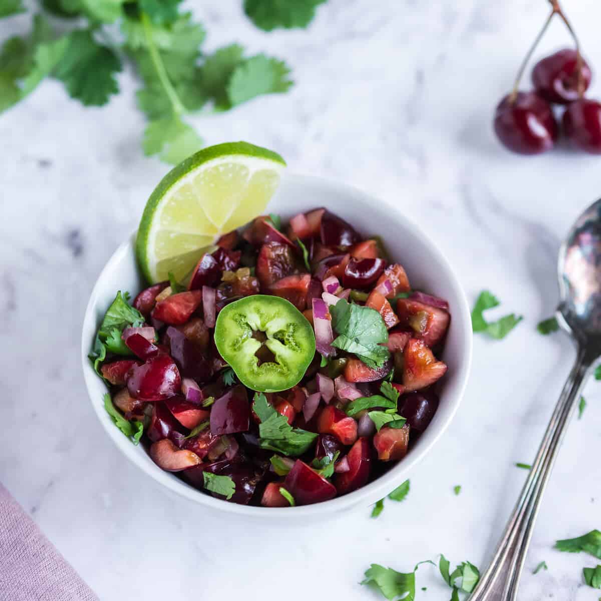 White bowl of fresh cherry salsa garnished with cilantro and a slice of jalapeno. Spoon alongside the bowl.