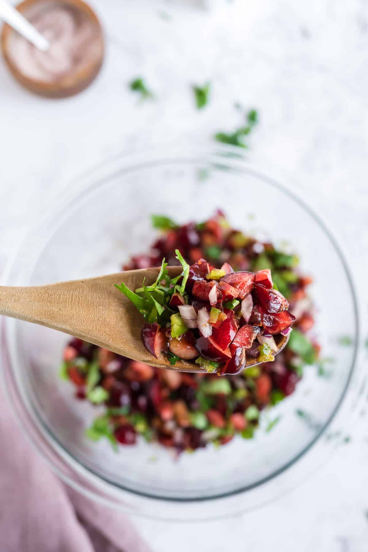 Cherry salsa on a wooden spoon being held above a bowl of salsa.