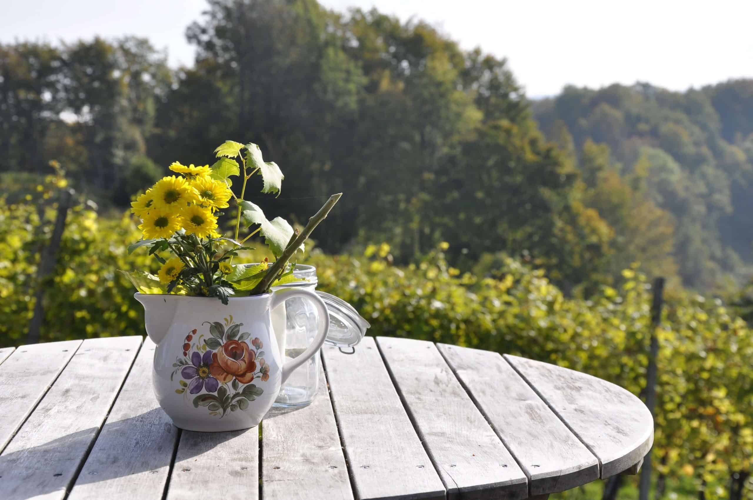 Rustic wood table outside with a vase of flowers on it. Surrounded by trees and nature.
