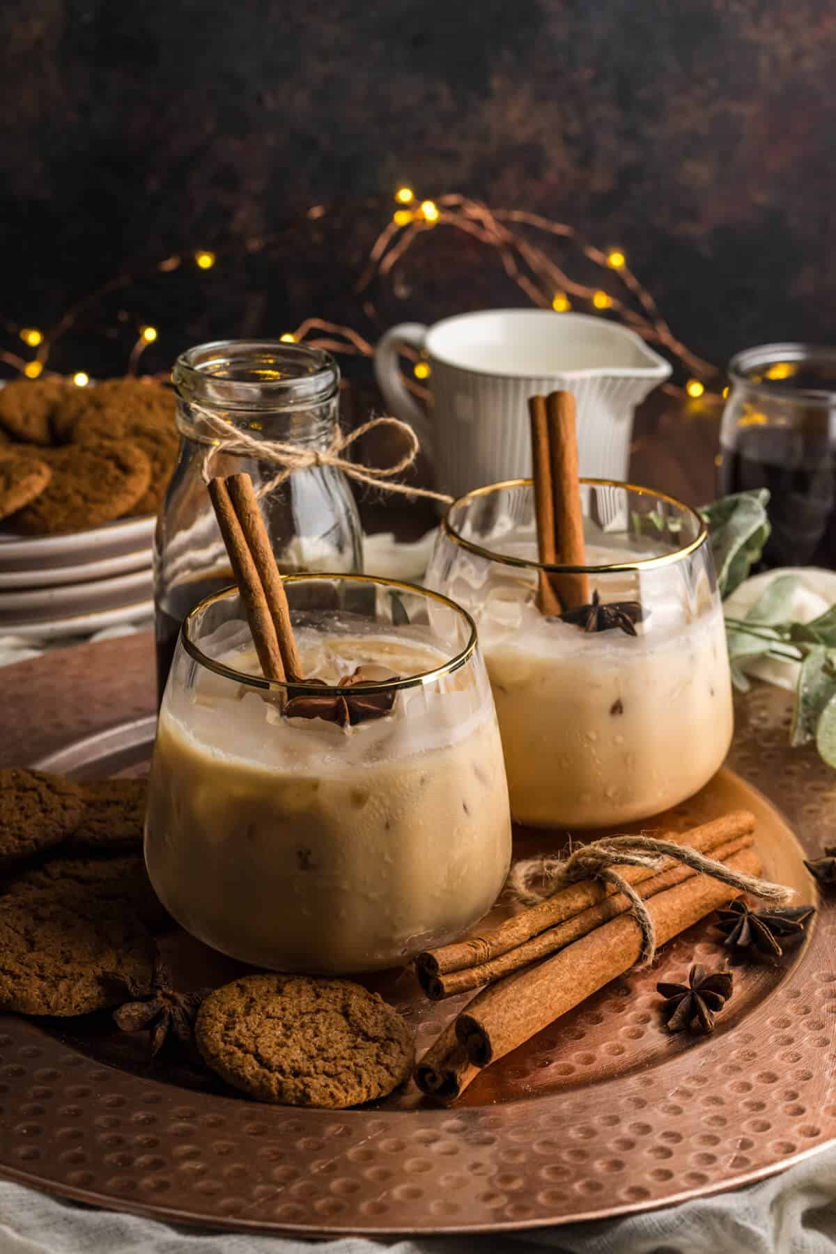 Two Gingerbread White Russians (non-alcoholic) in a gold rimmed ribbed stemless glass, garnished with cinnamon sticks and star anise. The drinks are surrounded with gingersnap cookies and more cinnamon sticks with twinkle lights in the background.