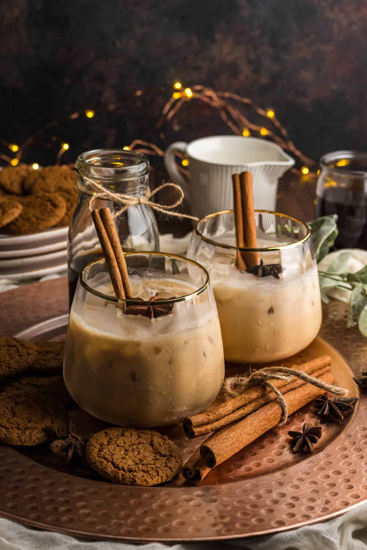 Two Gingerbread White Russians (non-alcoholic) in a gold rimmed ribbed stemless glass, garnished with cinnamon sticks and star anise. The drinks are surrounded with gingersnap cookies and more cinnamon sticks with twinkle lights in the background.