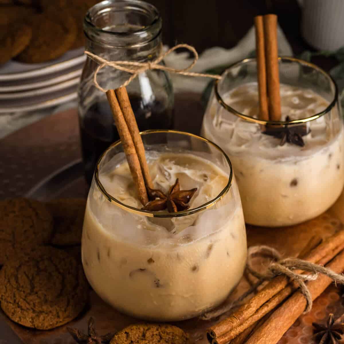 Two Gingerbread White Russians (non-alcoholic) in a gold rimmed ribbed stemless glass, garnished with cinnamon sticks and star anise. The drinks are surrounded with gingersnap cookies and more cinnamon sticks with twinkle lights in the background.