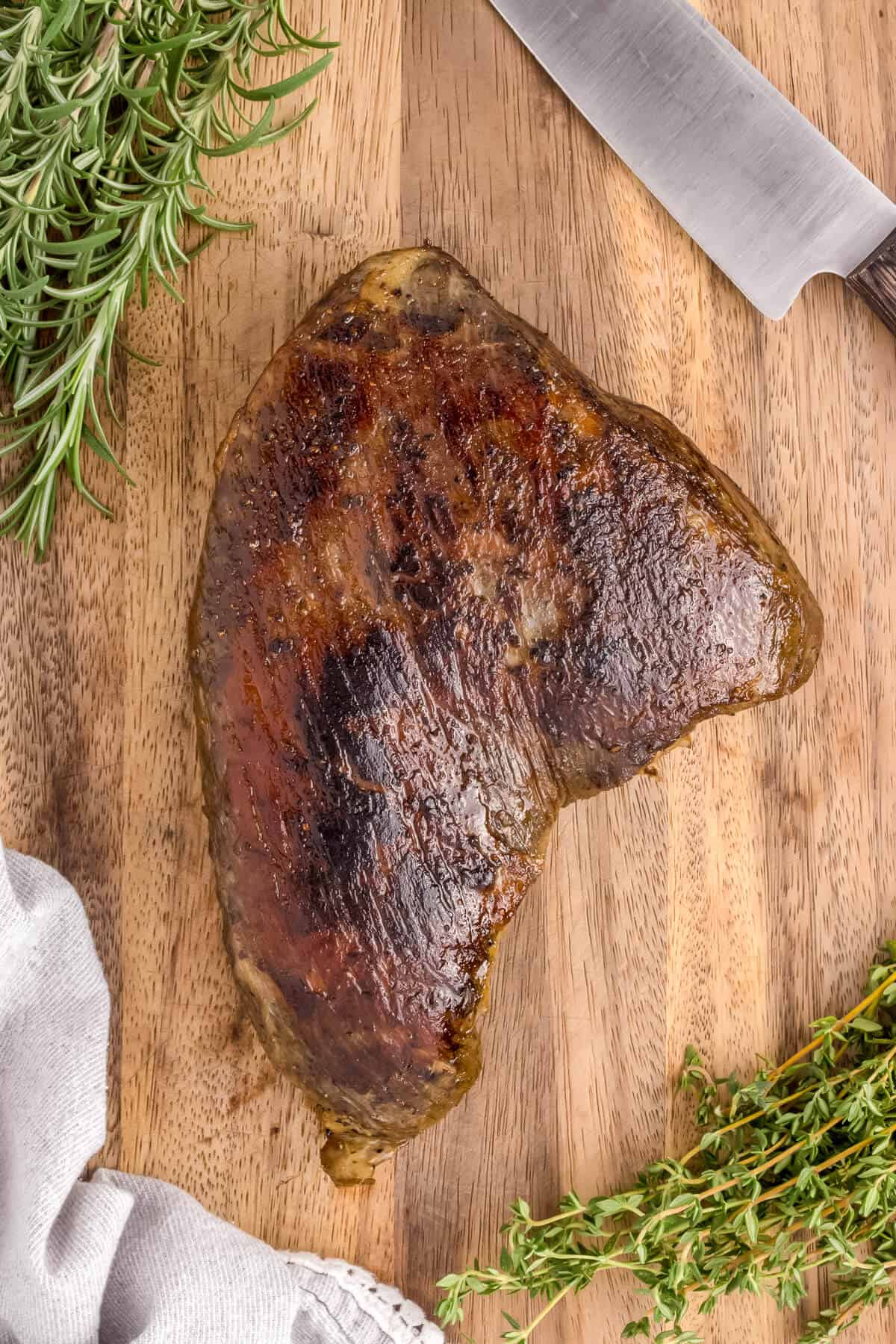 Just seared tri-tip roast on a cutting board surrounded by fresh rosemary and thyme.