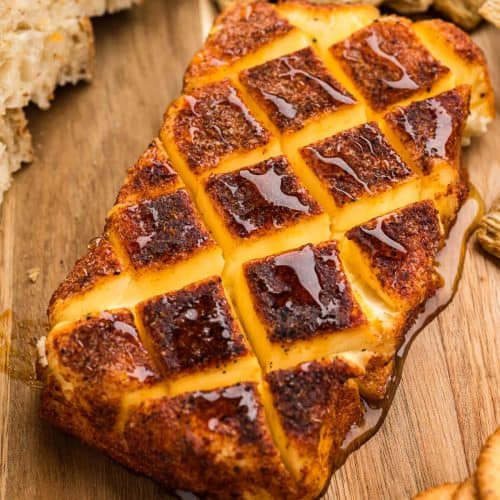 Smoked cream cheese with a cross hatch pattern and a drizzle of honey, surrounded by crackers and bread on a wooden platter.