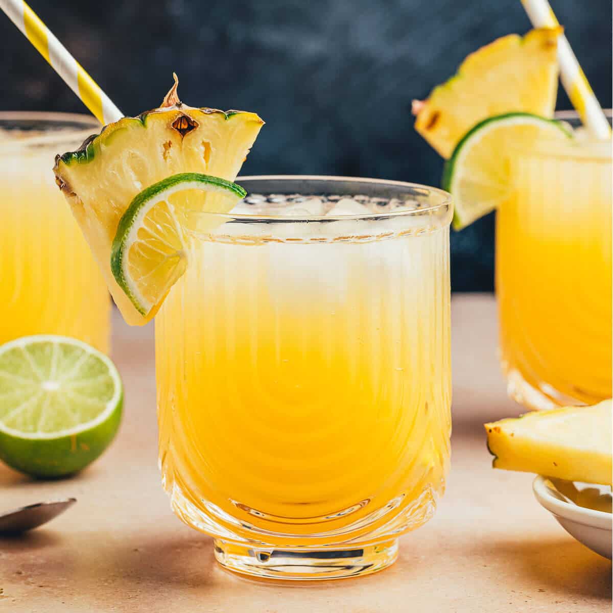 Pineapple ginger beer mocktails served in stunning crystal glassware with U shaped ribbing. Garnished with a pineapple wedge and a lime wedge, and a yellow and white striped straw.