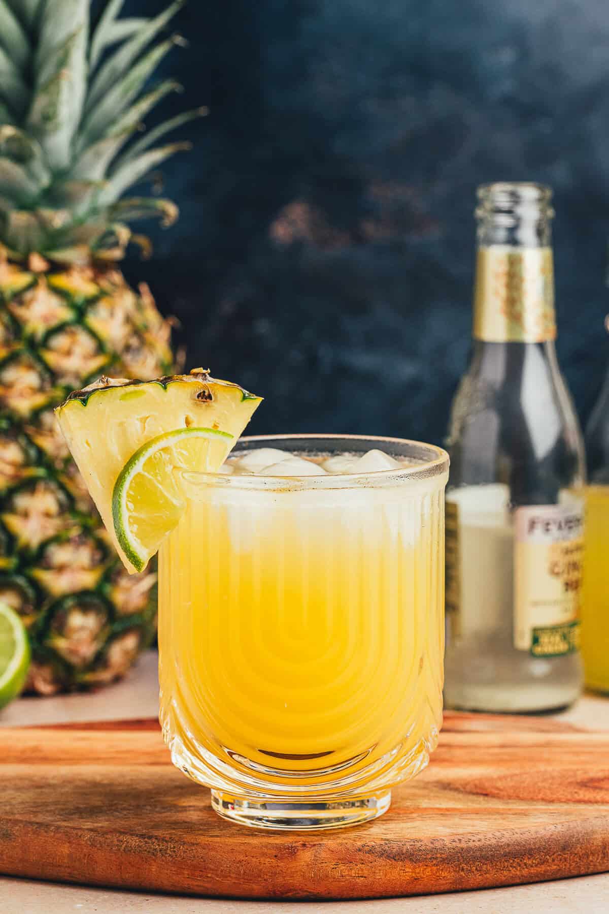 Glass of ginger beer with pineapple on a wooden serving board. A pineapple and empty bottle of gingerbeer stand in the background.
