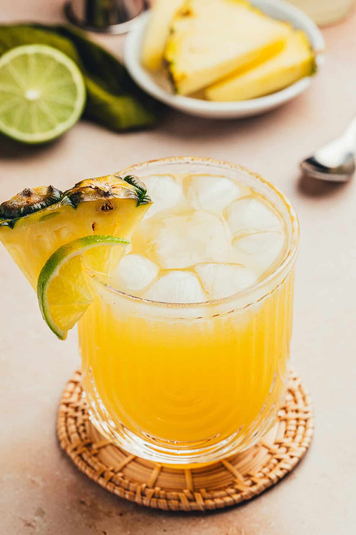 A pineapple ginger beer mocktail garnished with a slice of lime and pineapple wedge. Pineapple slices and lime slices are in the background.