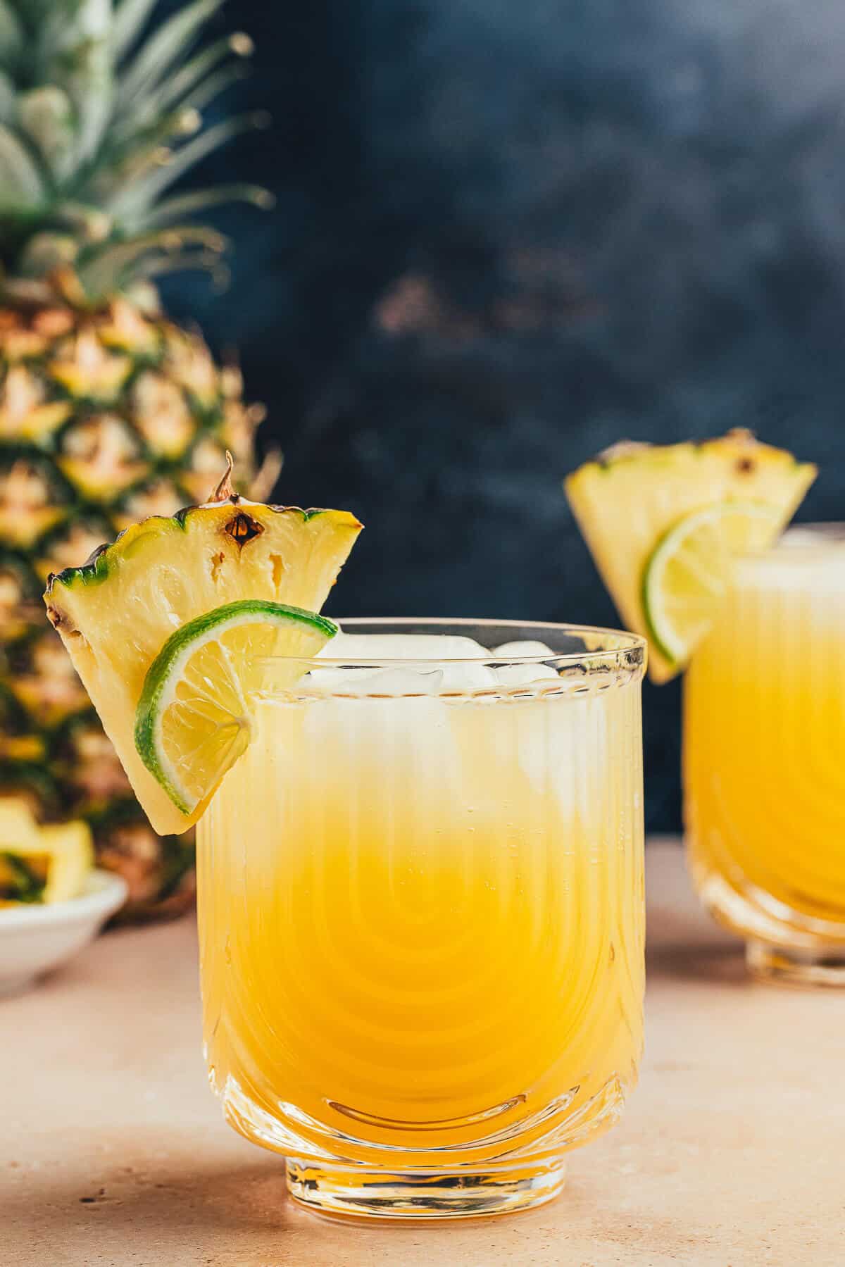 A glass of refreshing pineapple ginger beer mocktail garnished with a pineapple wedge and slice of lime. Another drink and a pineapple are in the background.