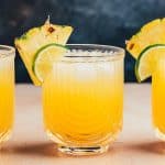 Three Pineapple mocktails served in crystal glasses with U shaped ribbing. The drinks are garnished with pineapple slices and lime slices.