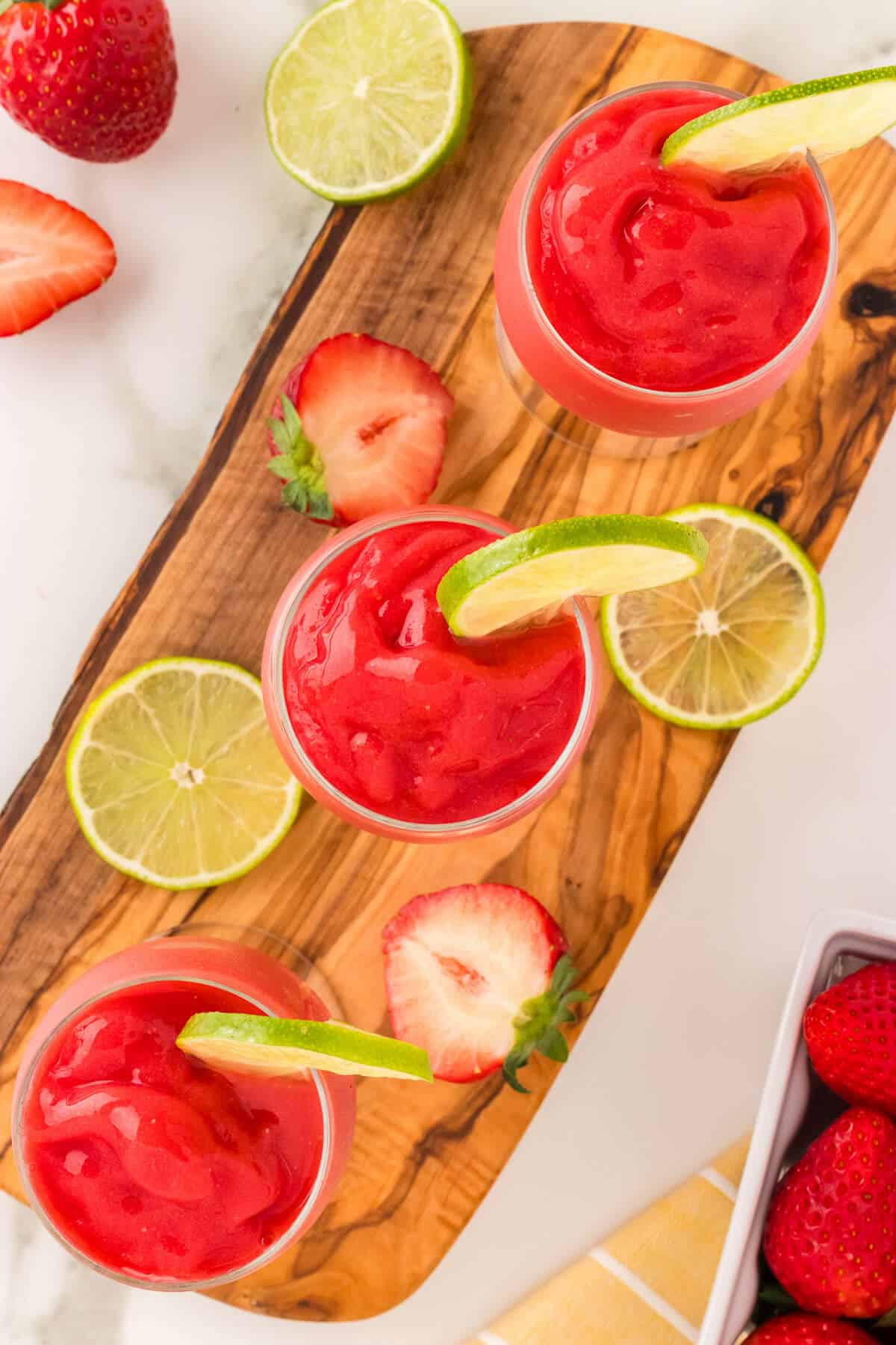 3 freshly made frozen virgin strawberry daiquiris on a wooden platter. Drinks are garnished with lime wheels, a few lime wheels and strawberries are scattered around them.