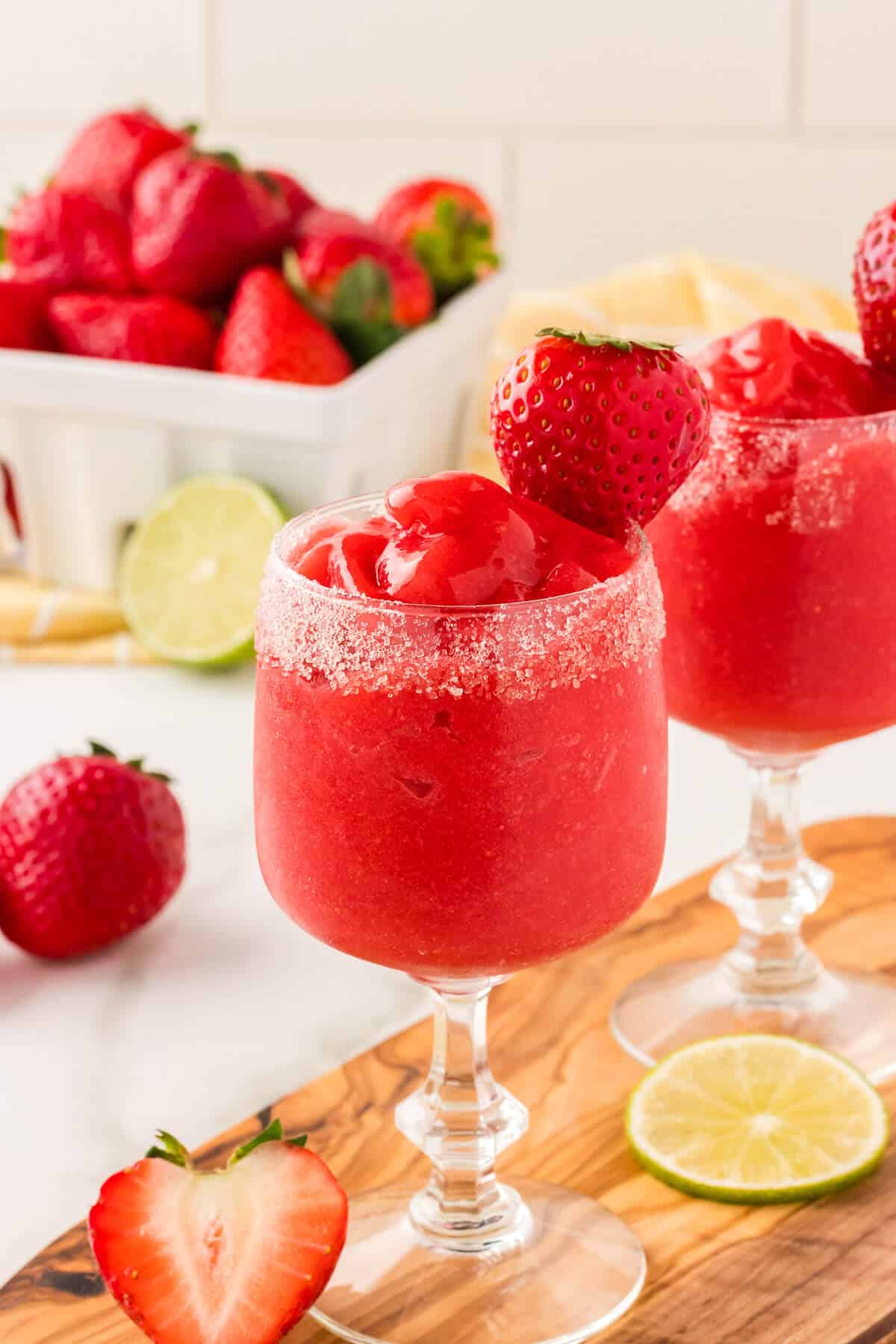 Virgin strawberry daiquiris in a sugar rimmed glass, garnished with a strawberry. A quart of strawberries is in the background, and there are a few strawberries and lime wheels scattered.