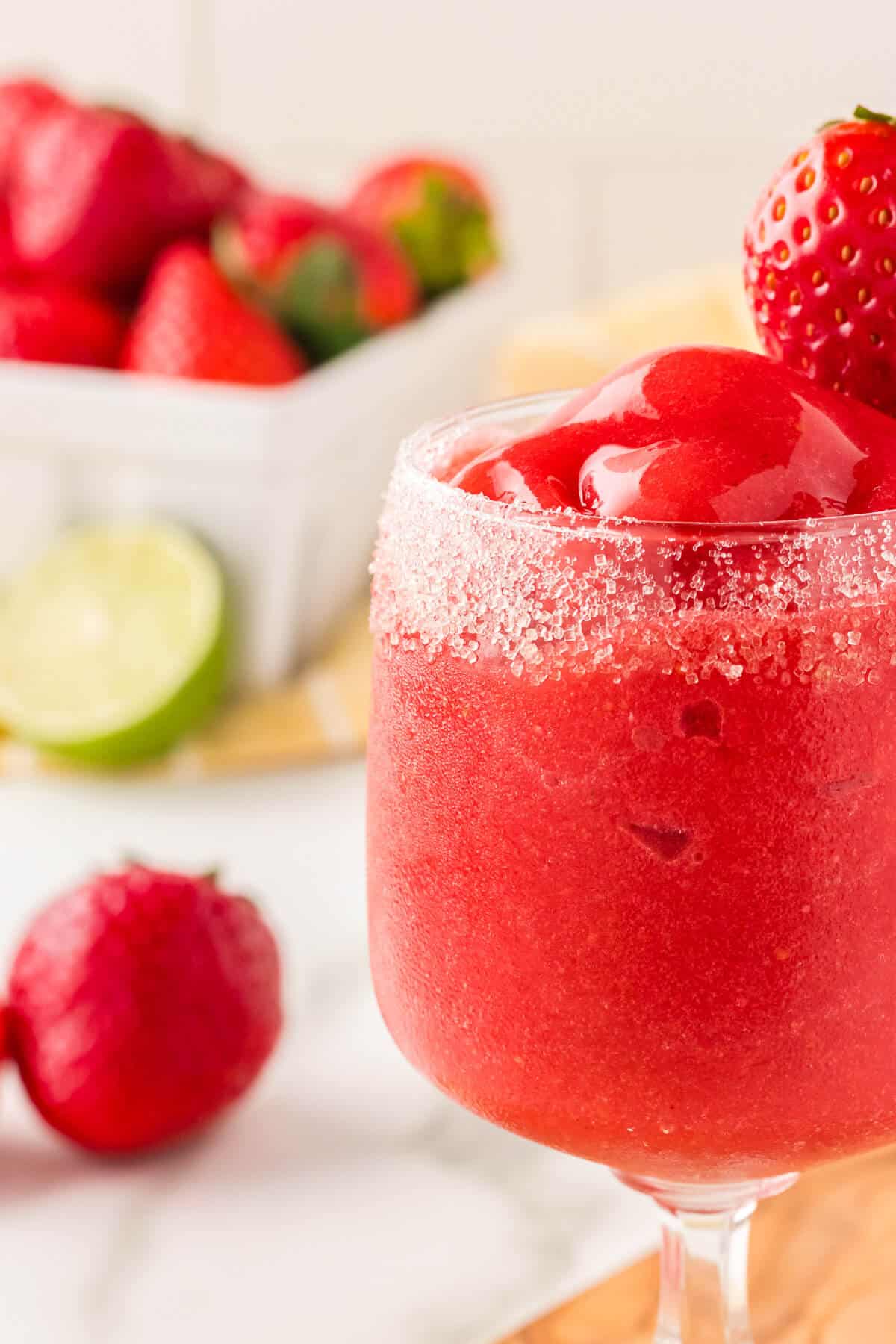 Nonalcoholic frozen strawberry daiquiri in a sugar rimmed glass with strawberries and limes in the background.