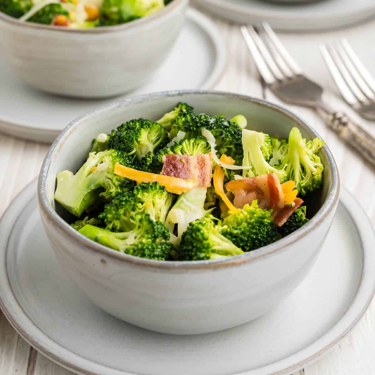 Serving of Chicken Salad Chick Broccoli Salad in a gray bowl, sitting on top of a small plate with forks in the background.