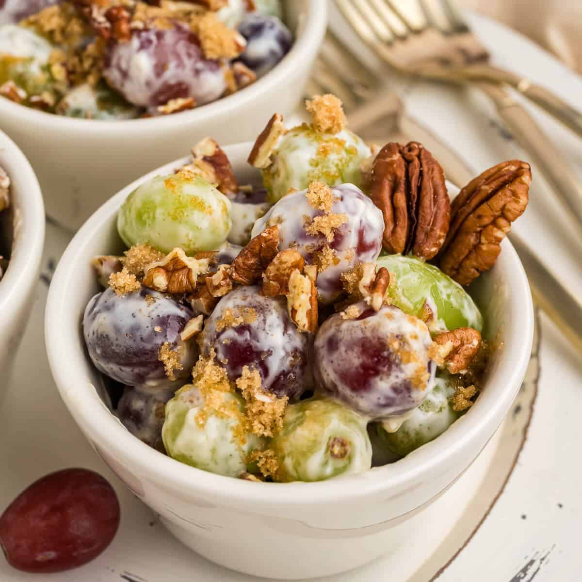 Close up picture of grape salad in a small serving bowl, garnished with brown sugar and chopped pecans. A couple of whole pecans in the corner of the bowl. There are more bowls of grape salad in the background and some gold forks.