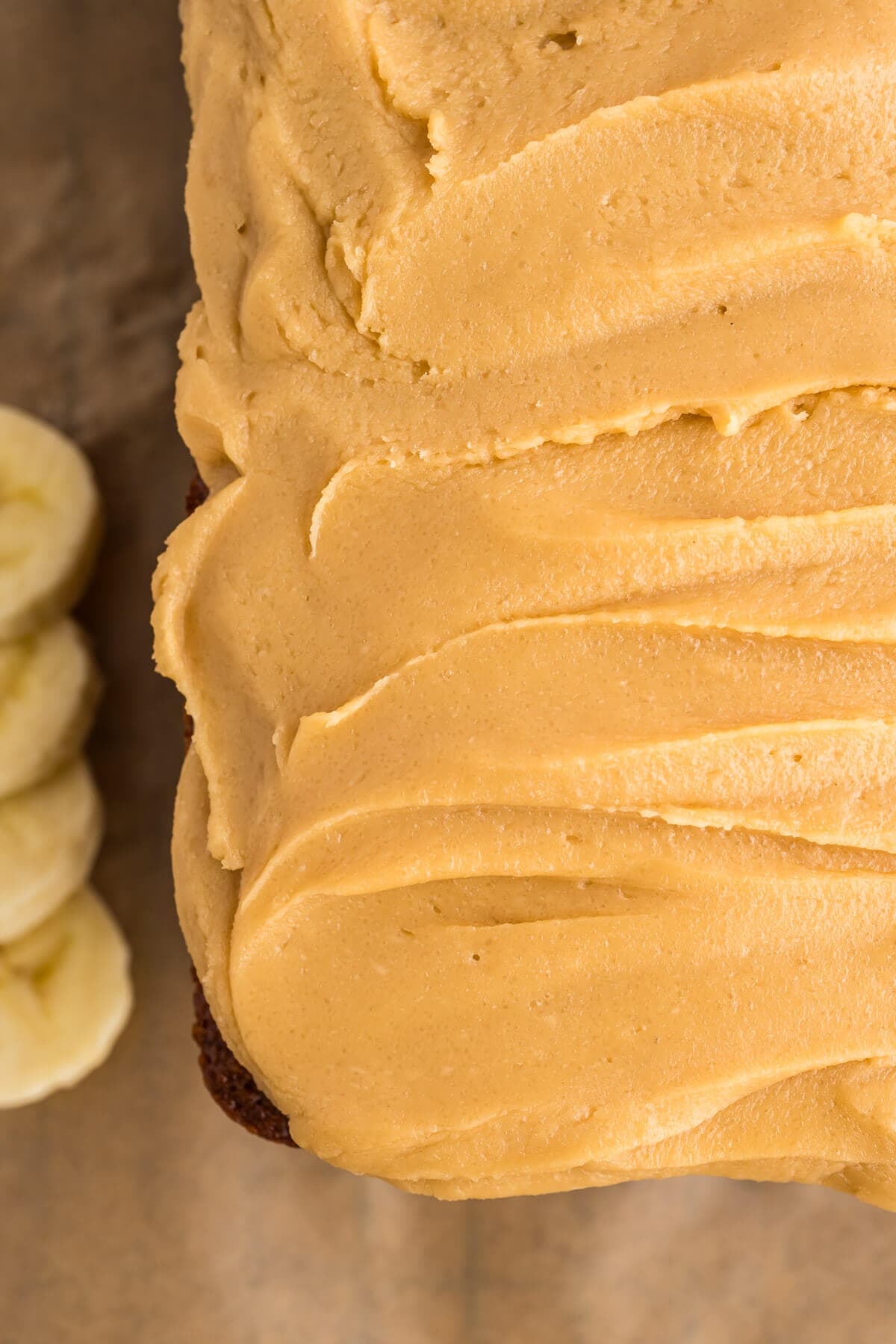 Close up overhead view of banana bread that has thickly spread caramel frosting. The loaf fills most of the frame with some sliced bananas peeking out on the left side next to the loaf.