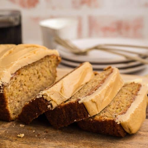 A loaf of banana bread with caramel icing, with three slices laying down on a cutting board next to the loaf itself.