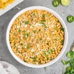 Elote dip in a round serving dish surrounded with chips, some lime slices and jalapeno slices, and some fresh cilantro.