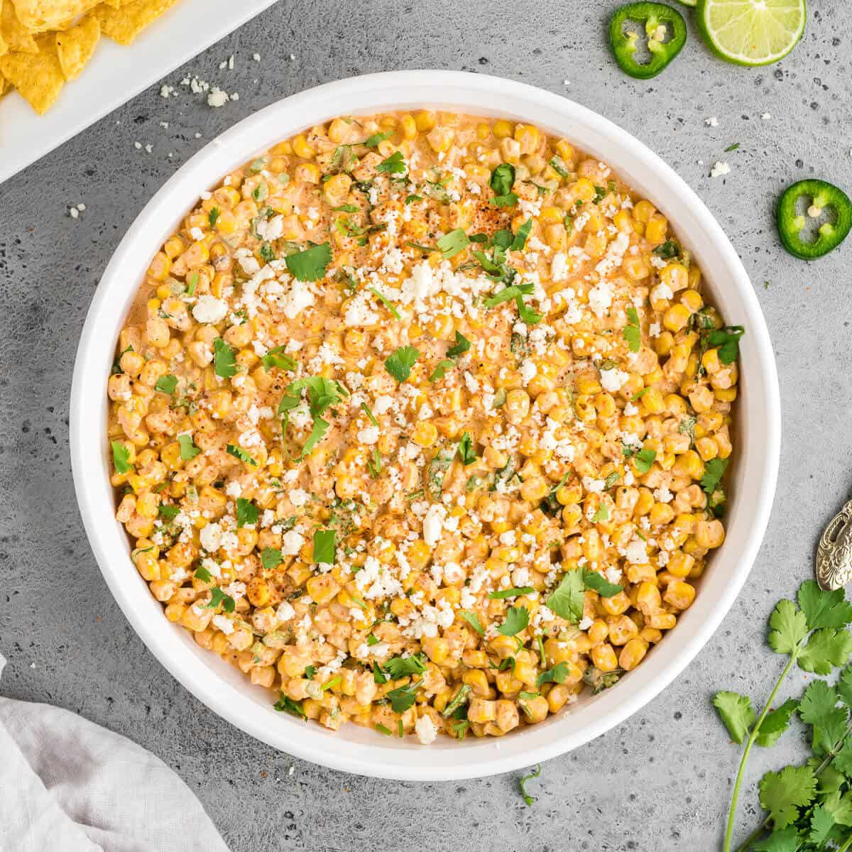Elote dip in a round serving dish surrounded with chips, some lime slices and jalapeno slices, and some fresh cilantro.