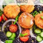 Crispy goat cheese topped on a bed of mixed greens, onions, sliced cucumbers, blackberries and cherry tomatoes. It's drizzled with Dijon Vinaigrette. Text overlay reads: Crispy Goat Cheese Salad.