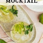 Overhead view of two cucumber mocktails on a beige oval tray. Drinks are garnished with fresh mint, and a cucumber ribbon on a gold cocktail spear. Fresh limes, mint, and cucumbers are around the drinks. Text overlay reads: Cucumber Lime Mocktail.