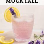Lavender Lemonade Mocktail in a clear glass that has a clear decorative design. The lemonade is pink in color and garnished with a lemon wheel and a lavender bud. There are a few lavender buds on the right of the glass and a couple slices of lemon to the left. The glass is on a round white wood board. Text overlay reads: Lavender Lemonade Mocktail.