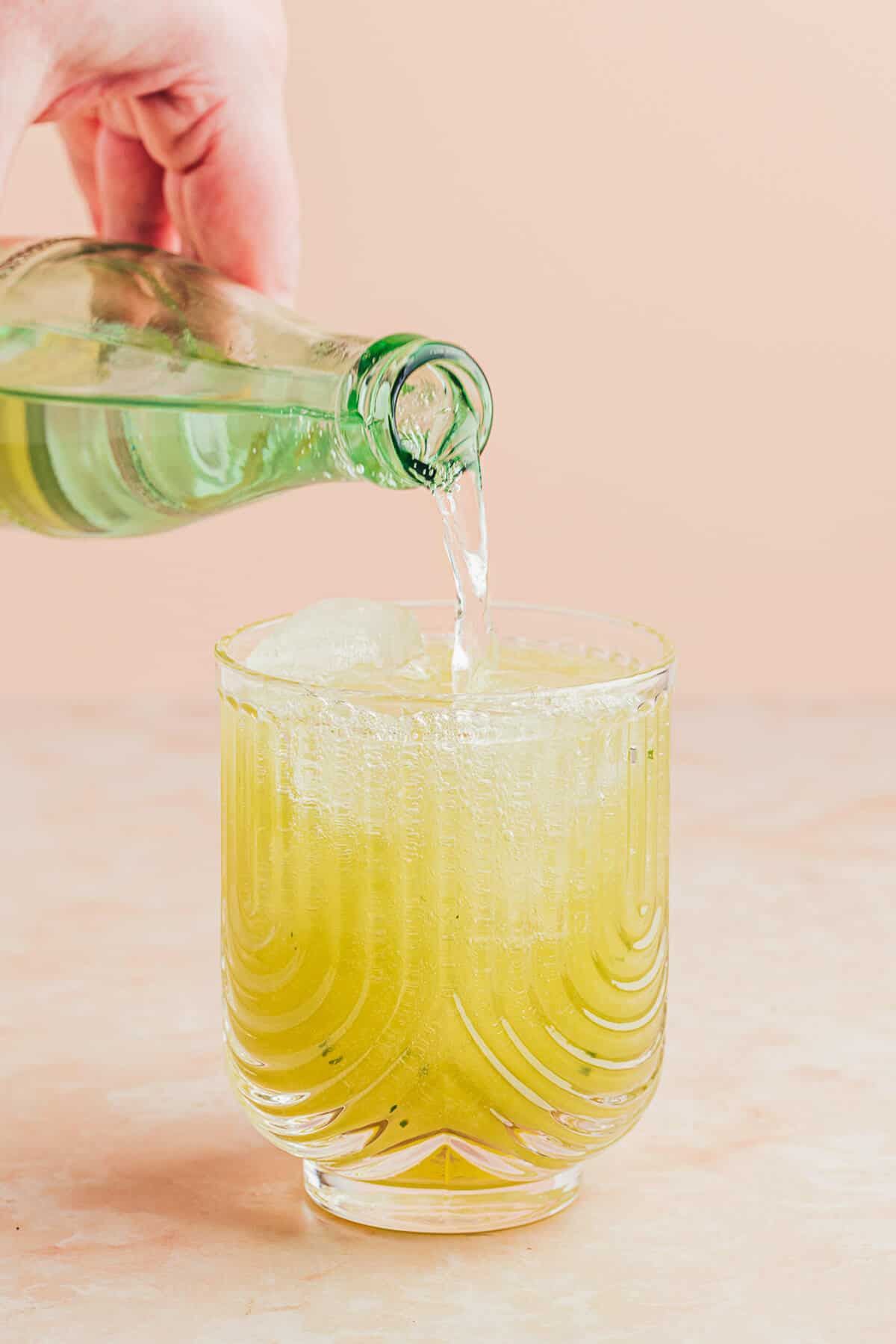 Sparkling water being poured to 'top off' the cucumber mocktail in a clear glass with u-shaped ribbing.
