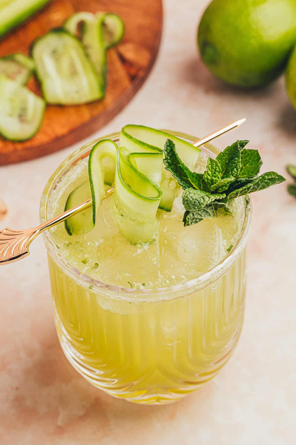 Cucumber lime Mocktail in a clear ribbed glass garnished with fresh mint and cucumber ribbon on a gold cocktail pick. Fresh whole limes and cucumber ribbons in the background.