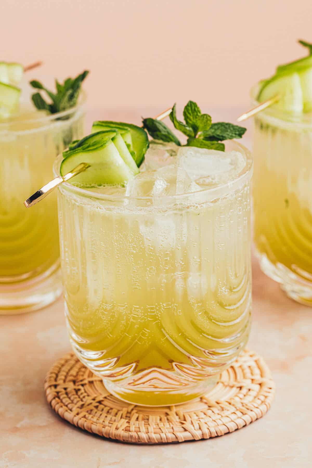 Cucumber lime Mocktail in a clear U-shaped ribbed glass garnished with fresh mint and cucumber ribbon on a gold cocktail pick. Two more drinks are in the background.