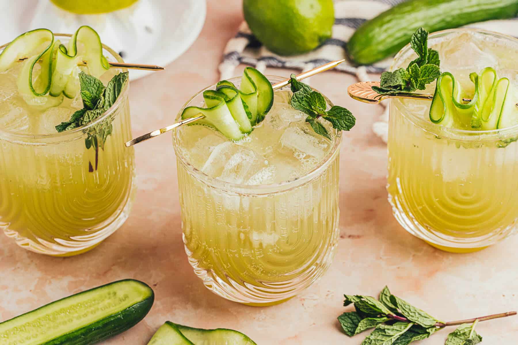 Three cucumber mocktails in a row. The drinks are in U-shaped ribbed clear glasses. There are fresh limes, cucumbers and mint leaves in the background and foreground.