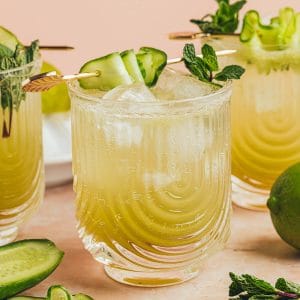 Cucumber lime Mocktail in a clear U-shaped ribbed glass garnished with fresh mint and cucumber ribbon on a gold cocktail pick. Two more drinks are in the background.