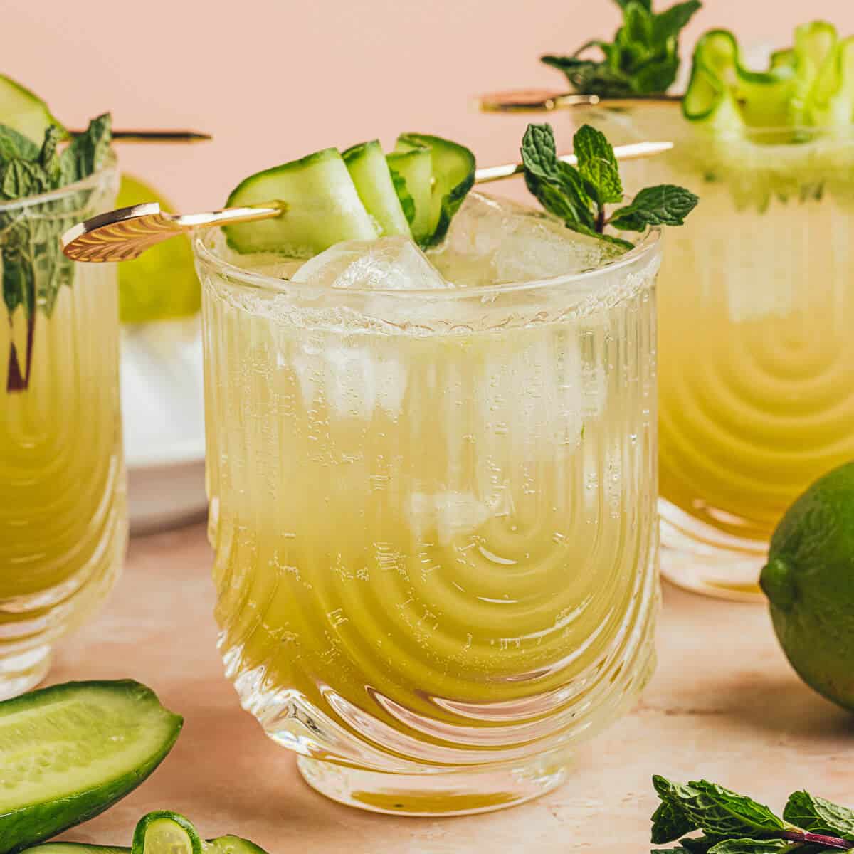 Cucumber lime Mocktail in a clear U-shaped ribbed glass garnished with fresh mint and cucumber ribbon on a gold cocktail pick. Two more drinks are in the background.