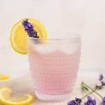 Lavender Lemonade Mocktail in a clear glass that has a clear decorative design. The lemonade is pink in color and garnished with a lemon wheel and a lavender bud. There are a few lavender buds on the right of the glass and a couple slices of lemon to the left. The glass is on a round white wood board.