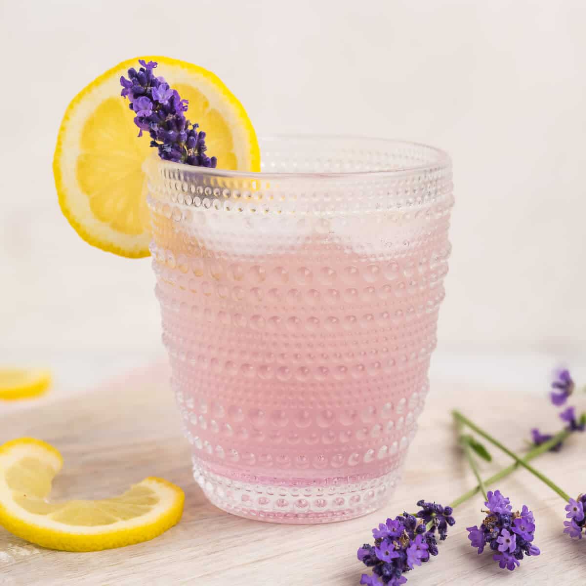 Lavender Lemonade Mocktail in a clear glass that has a clear decorative design. The lemonade is pink in color and garnished with a lemon wheel and a lavender bud. There are a few lavender buds on the right of the glass and a couple slices of lemon to the left. The glass is on a round white wood board.