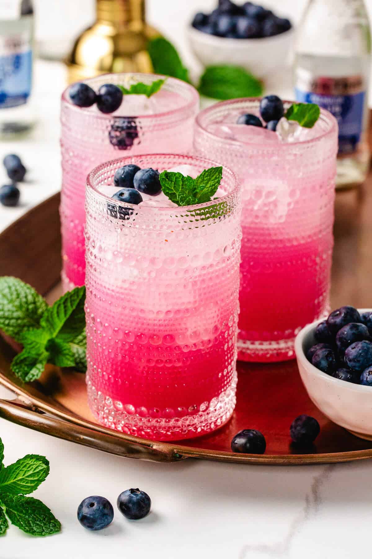 Three glasses of blueberry mint mocktail on a copper tray, garnished with fresh blueberries and mint leaves. The layered pink drink is served over ice in textured glasses, surrounded by extra blueberries and fresh mint on a white surface.