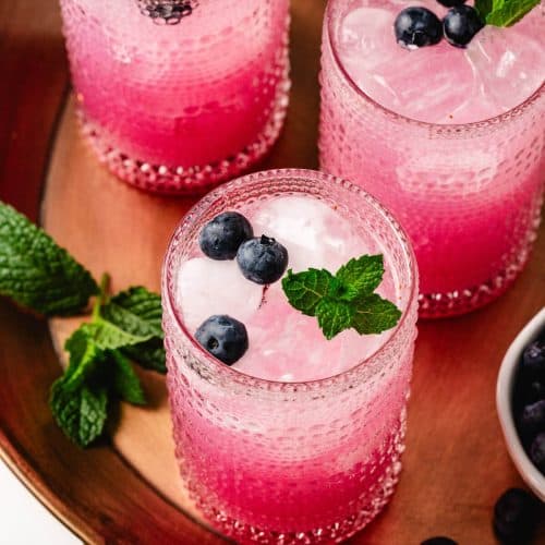 Top-down view of three blueberry mint mocktails served over ice in clear textured glasses, each garnished with fresh blueberries and mint sprigs. The drinks are presented on a copper tray with extra mint and blueberries.