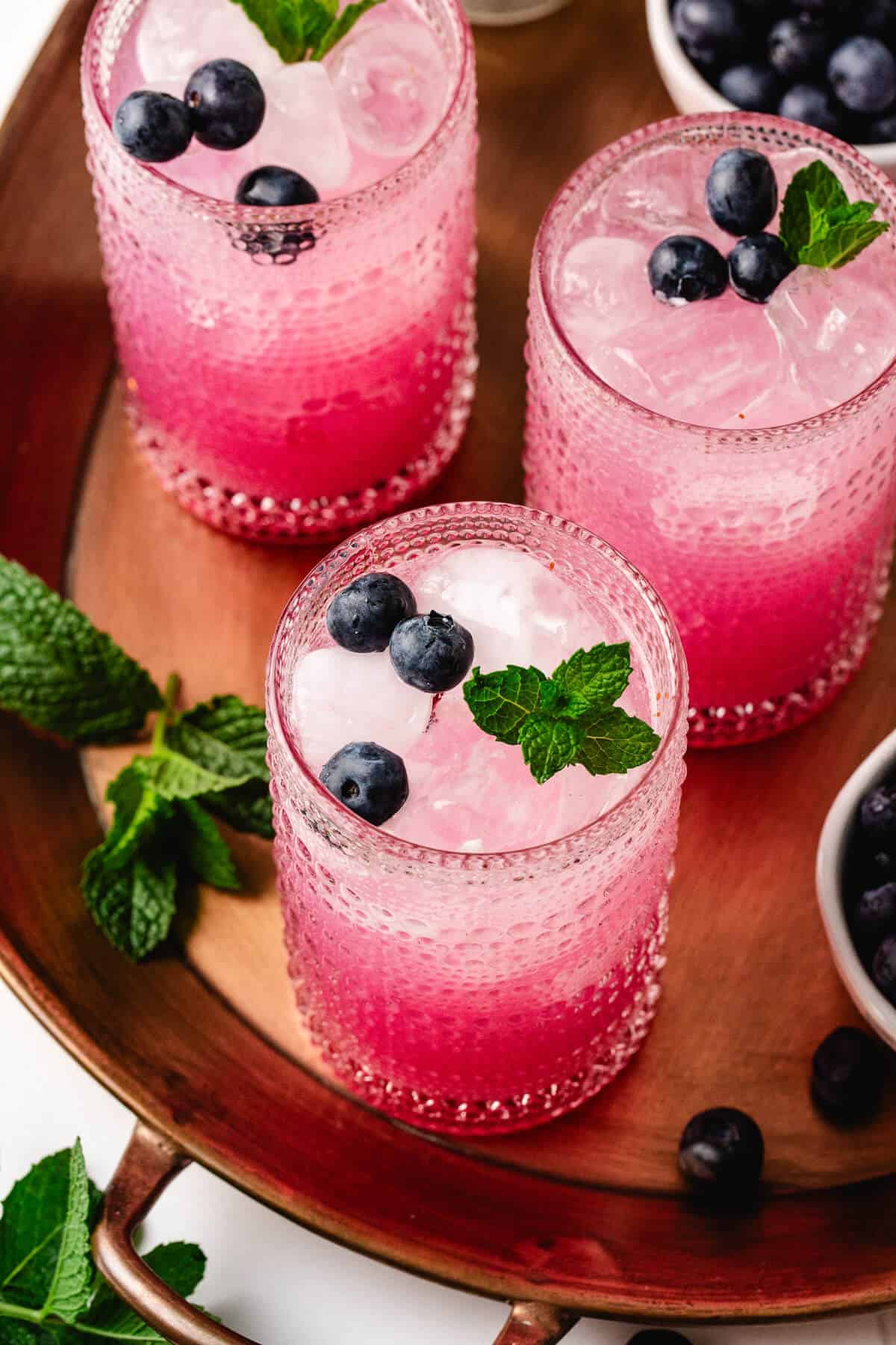 Top-down view of three blueberry mint mocktails served over ice in clear textured glasses, each garnished with fresh blueberries and mint sprigs. The drinks are presented on a copper tray with extra mint and blueberries.
