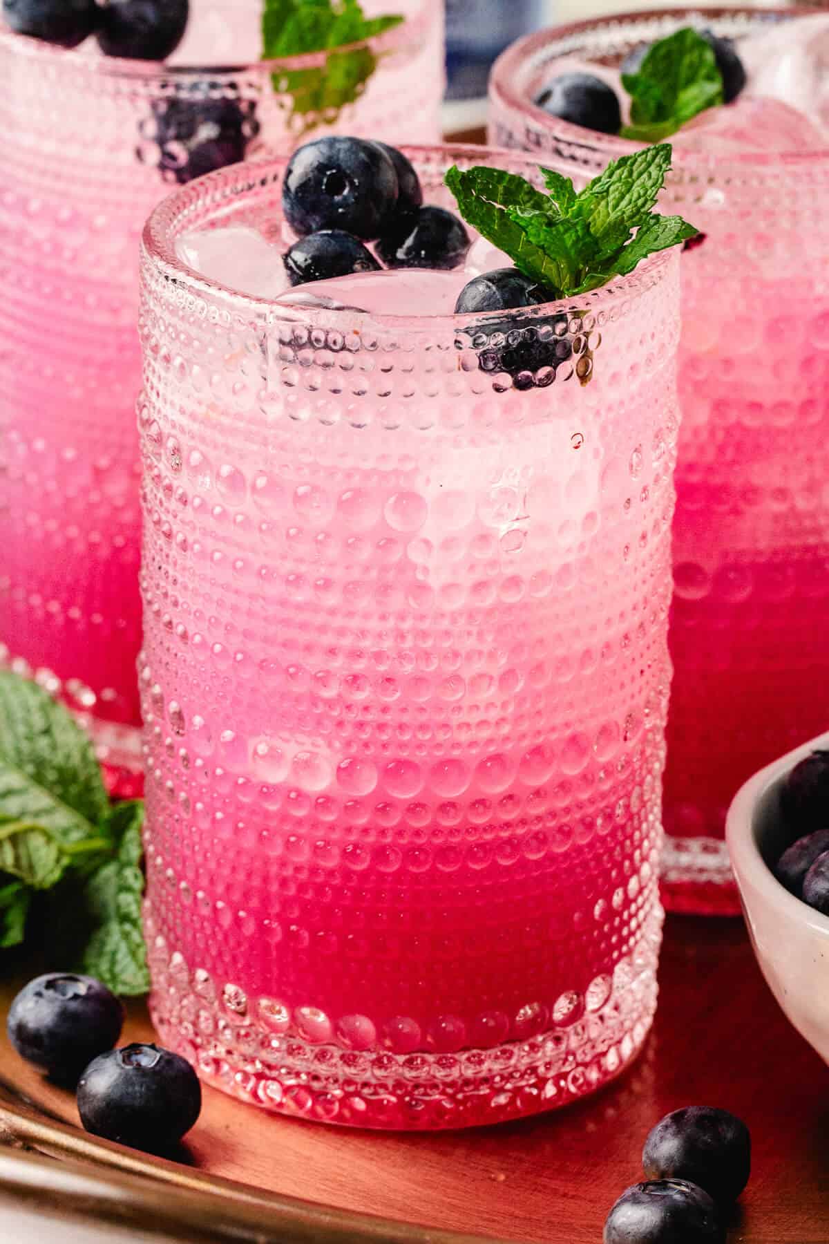 A close-up of a single blueberry mint mocktail in a clear beaded glass, showing off the ombré pink hue and icy top with blueberries and mint. Fresh fruit and herbs rest nearby on the copper tray.