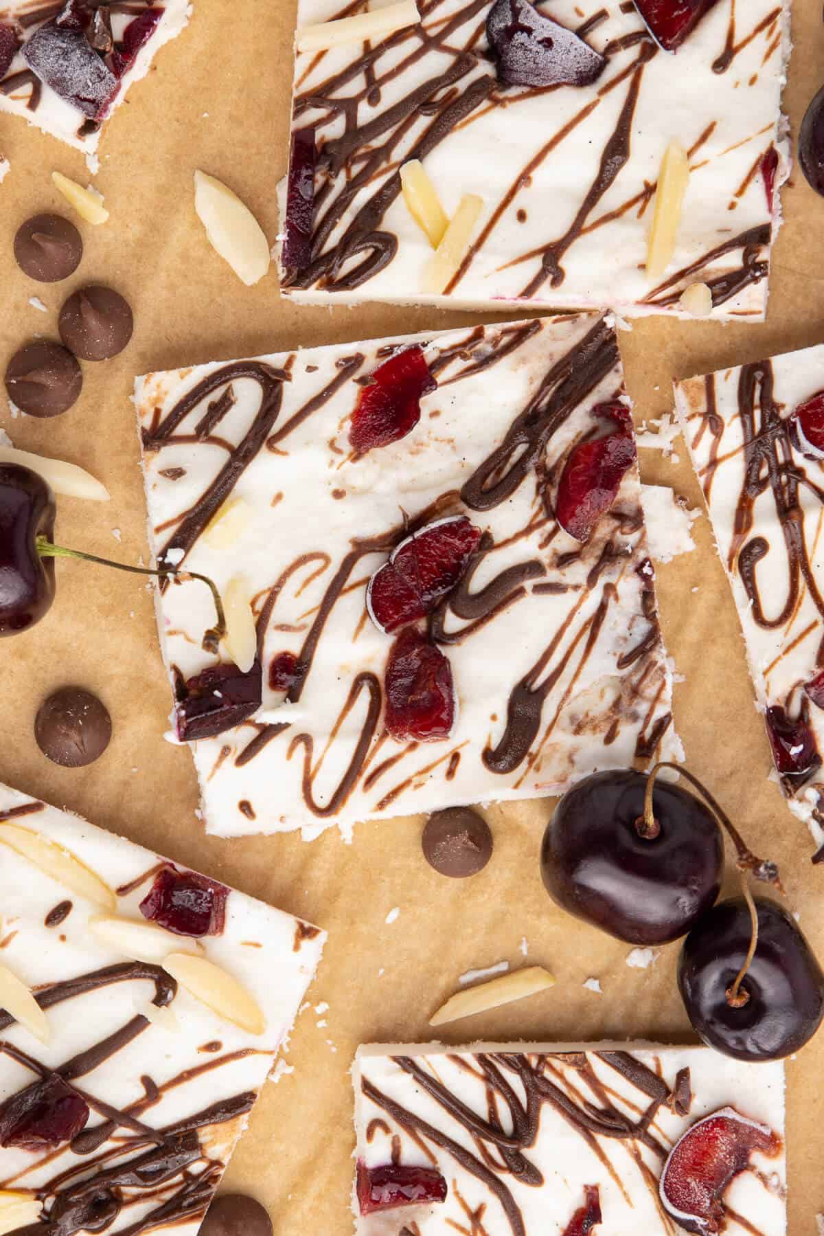 Several squares of Cottage Cheese Ice Cream Bark. The bark is drizzled with chocolate and has chopped cherries and slivered almonds in it. There are a few cherries, chocolate chips. and slivered almonds scattered around the pieces.