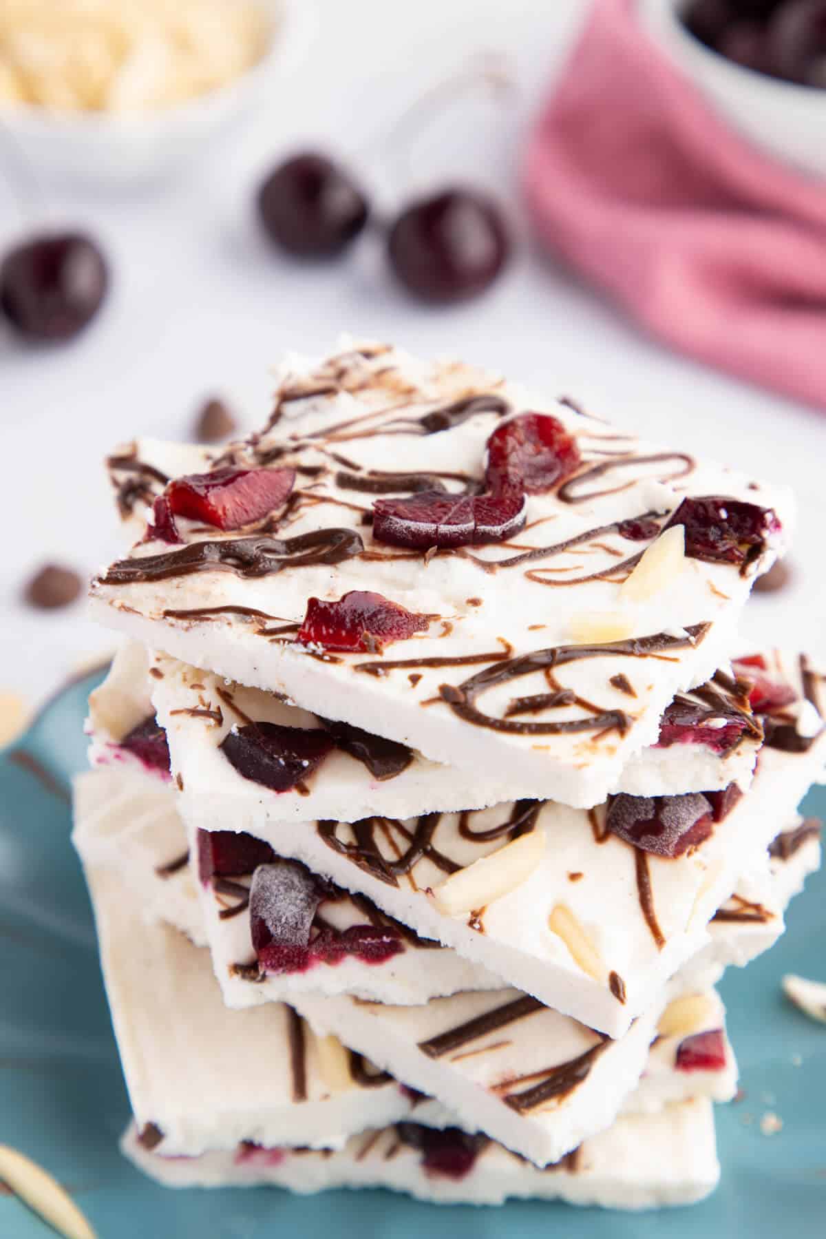 Stack of Cherry Chocolate Ice Cream Bark Squares on a plate. A few cherries and chocolate chips are in the background.