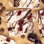 Several squares of Cottage Cheese Ice Cream Bark. The bark is drizzled with chocolate and has chopped cherries and slivered almonds in it. There are a few cherries, chocolate chips. and slivered almonds scattered around the pieces.