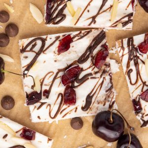 Several squares of Cottage Cheese Ice Cream Bark. The bark is drizzled with chocolate and has chopped cherries and slivered almonds in it. There are a few cherries, chocolate chips. and slivered almonds scattered around the pieces.