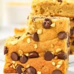 Peanut butter cookie bar standing on its side with a bite taken out, leaning against a stack of bars, topped with chocolate chips and chopped peanuts.
